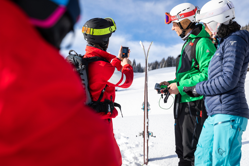 Un guide en combinaison rouge montre l'utilisation d'un ARVA à deux skieurs en montagne, lors d'une session de ski de randonnée sur la neige.