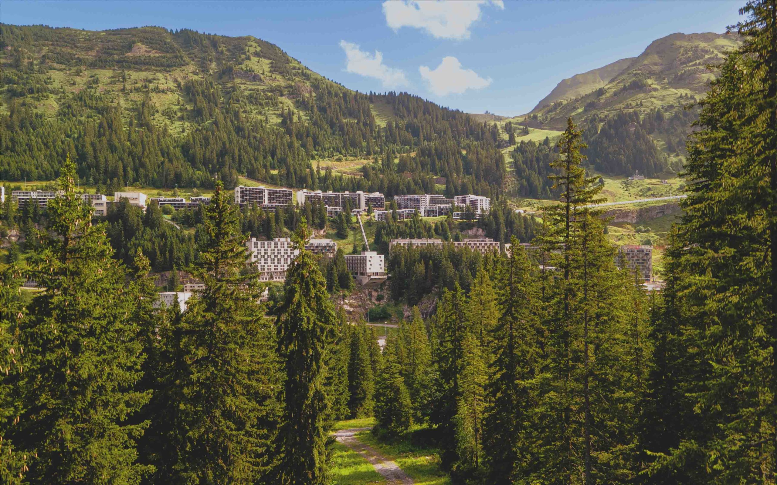 Panorama estival de la station de Flaine entourée de sapins et de montagnes, sous un ciel bleu.