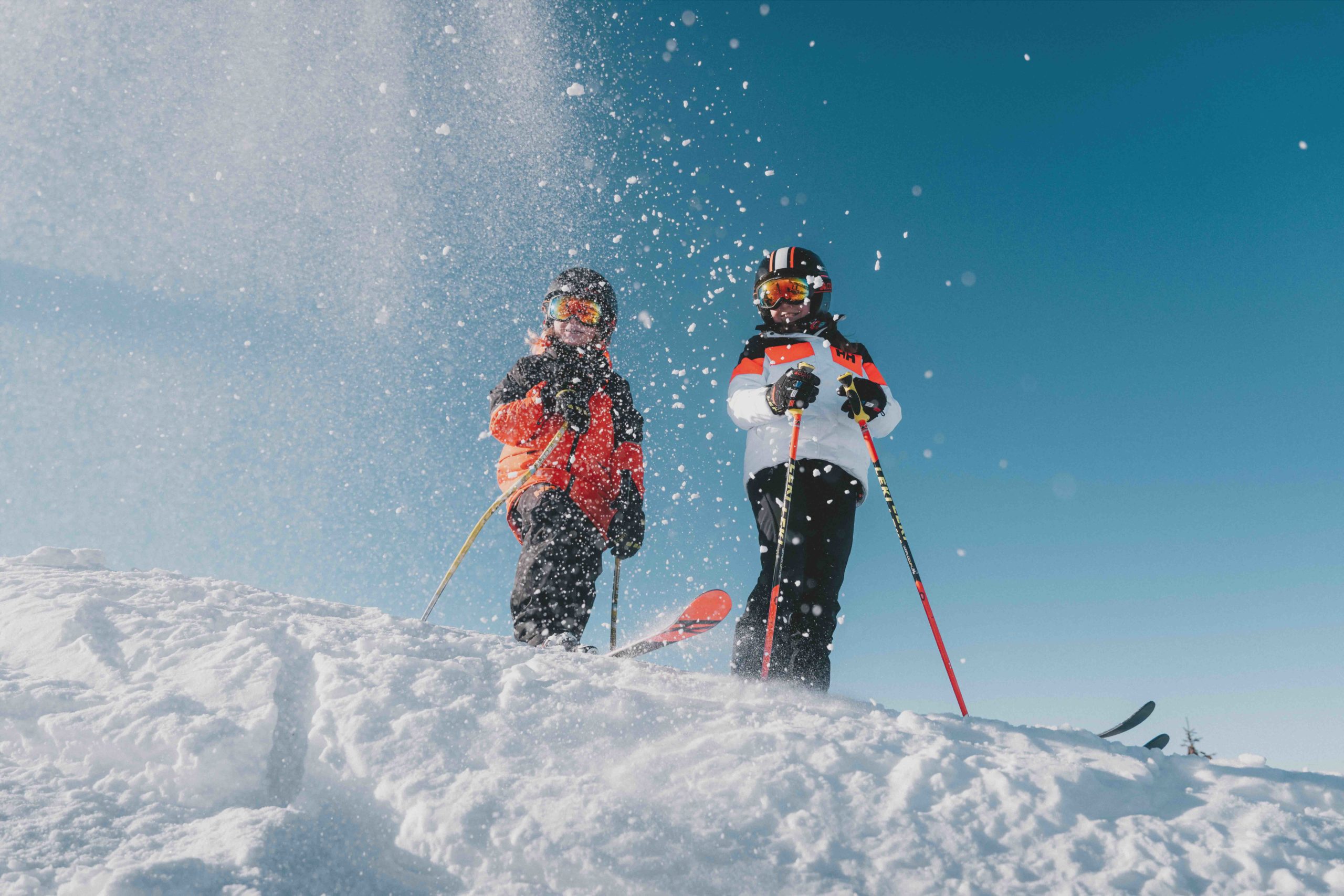 Deux enfants skieurs en combinaisons colorées sur une piste enneigée avec un ciel bleu en arrière-plan, à Flaine en Haute-Savoie.
